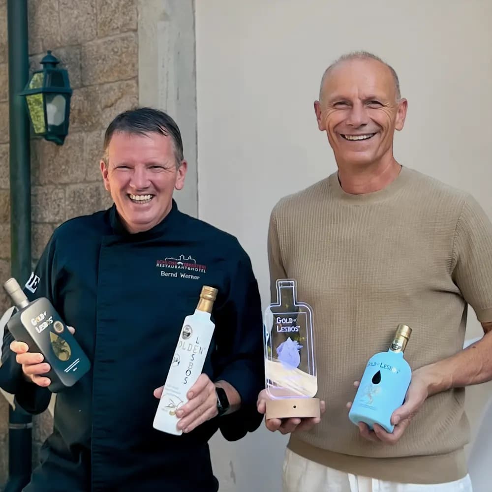 Küchenchef Bernd Werner und Produzent Anastasios Fortomaris halten mehrere Gold-of-Lesbos-Flaschen und eine Auszeichnung in den Händen